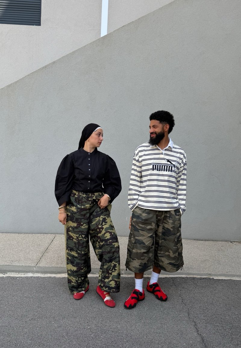 Deux personnes portant un pantalon camouflage et des chaussures rouges se tiennent dans une rue devant un mur gris, se faisant face et souriant.