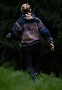 Mikina z flísu s vícebarevnými pruhy v odstínech fialové, modré a béžové. Má velkou kapuci a volný střih. Kombinována s černými legínami.