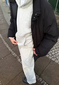 Sweat à capuche gris avec poche avant, associé à un pantalon de jogging assorti. Veste matelassée noire, texture unie, et col rond. Chaussures noires à enfiler.