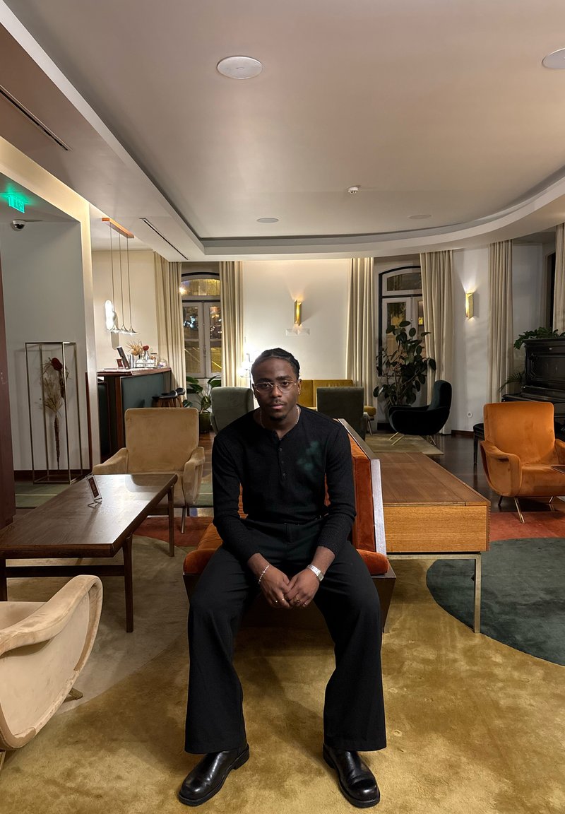 Mees, kes kannab musta, istub oranžil pingil moodsas elutoas, kus on erinevaid toole, laudu, taimi ja sooja valgustust.