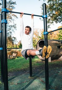 Juodas metalinis traukimosi strypas su mėlynais sujungimais, žmogus kabantis, dėvintis baltą ilgarankovį marškinį ir pilkas šortus, juodas sportines batus su geltonais padais.