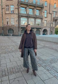 Brun trenchcoat over en svart hettegenser, kombinert med vide, falmede bukser og brune ankelstøvler. Brosteinsbelagt fortau og bakgrunn av mursteinsbygning.