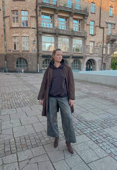 Abrigo trench marrón sobre una sudadera negra, combinado con pantalones de pierna ancha y descoloridos, y botas de tobillo marrones. Fondo de pavimento de adoquines y edificio de ladrillo.