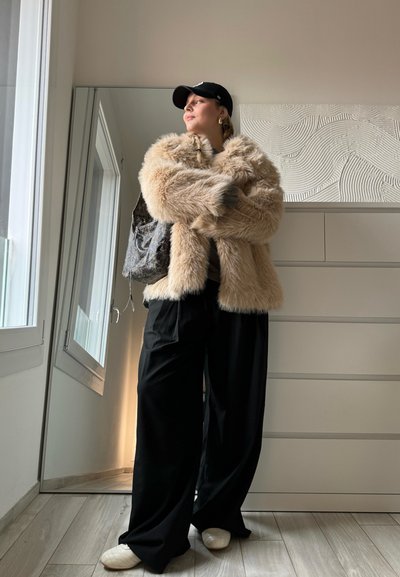 Chaqueta de pelo sintético en color beige claro, pantalones anchos negros y zapatos blancos. El fondo presenta un armario con espejo y una pared texturizada.