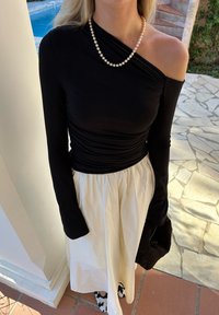 Top preto de manga longa com um decote assimétrico, combinado com uma saia branca fluída, e acentuado por um colar de pérolas e uma mala preta.
