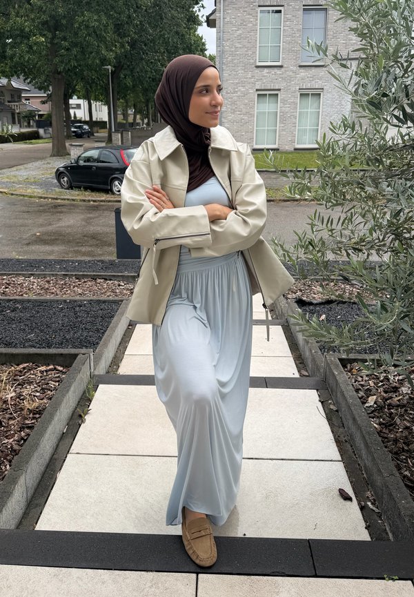 Vrouw draagt een lichtblauwe jurk, beige jas, bruine hijab en bruine loafers en loopt op een geplaveid pad naast planten en woongebouwen.