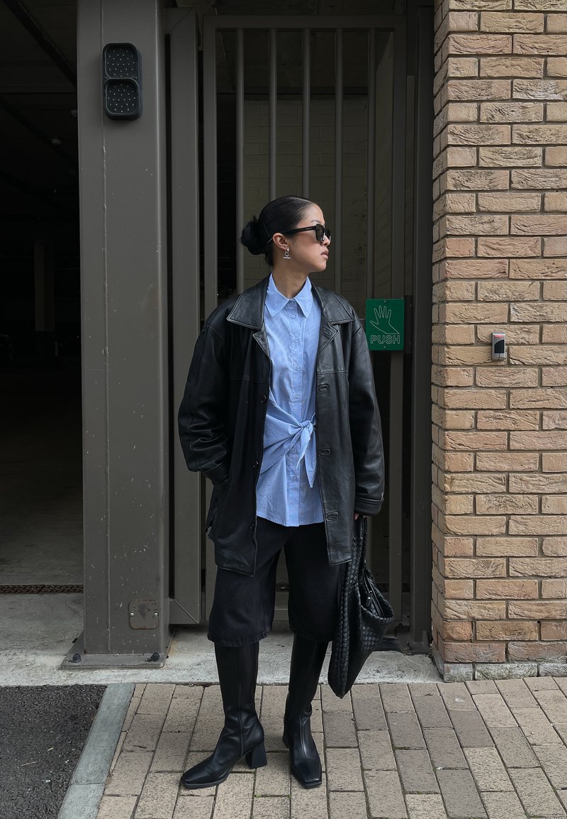 Frau mit Sonnenbrille, die eine schwarze Lederjacke, blaues Hemd, schwarze Hose und Stiefel trägt, steht bei einem Tor mit einem "drücken"-Schild.