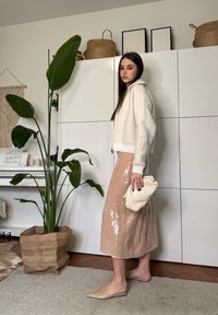 Νεαρή γυναίκα με μπεζ φούστα με παγιέτες, λευκό χνουδωτό σακάκι και γυναικεία nude παπούτσια, κρατώντας κρεμ τσάντα χειρός, στέκεται δίπλα σε ψηλό πράσινο φυτό σε πλεκτό καλάθι.