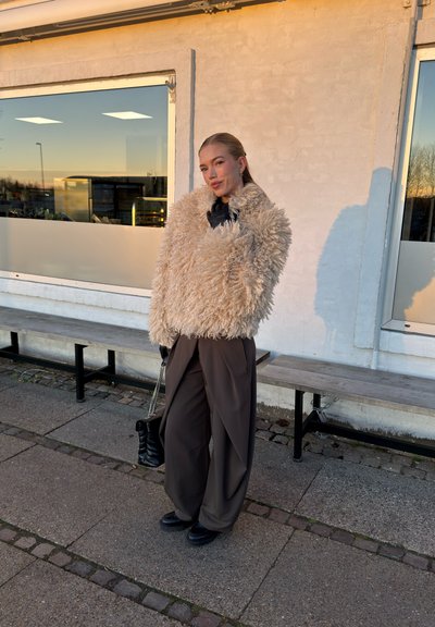 Chaqueta de piel sintética beige, camisa negra, pantalones marrones sueltos y zapatos de plataforma negros. Sostiene un bolso negro, con una pared gris claro detrás.