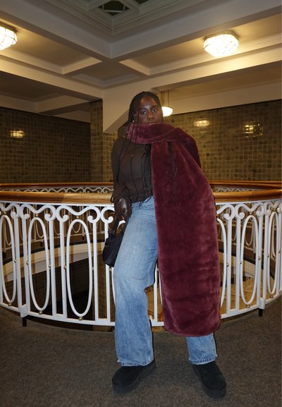 Mujer con largas trenzas que lleva un abrigo de piel sintética burdeos sobre una blusa marrón y vaqueros, de pie en el interior junto a una barandilla blanca decorativa.