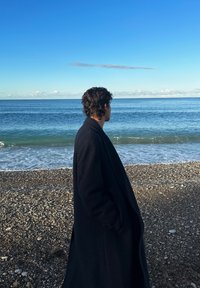 Long manteau noir, personne debout faisant face à l'océan. La texture est lisse contre la plage de galets, avec une eau bleue calme sous un ciel clair.