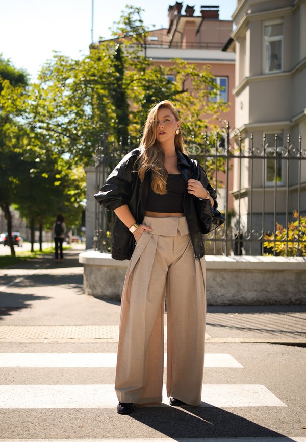 Femme aux longs cheveux portant un crop top noir, une veste noire, un pantalon beige à jambes larges et des chaussures noires, debout sur un passage piéton en ville.