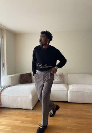 Hombre con suéter negro y pantalones grises de pie junto a un sofá blanco con cojines estampados en una sala de estar luminosa.