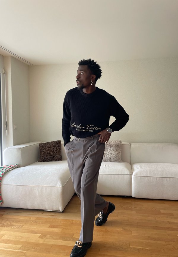 Man wearing black sweater and gray pants stands beside white sofa with patterned pillows in bright living room.