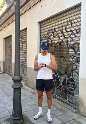 Figura masculina atlética vestida con una camiseta sin mangas blanca y pantalones cortos negros, revisando un smartwatch. Zapatillas brillantes con detalles en rosa. Fondo de grafitti.