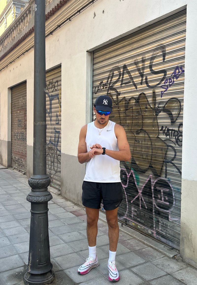 Figura masculina atlética vestindo uma camiseta sem mangas branca e calções pretos, a verificar um smartwatch. Ténis brilhantes com detalhes em rosa. Fundo de graffiti.