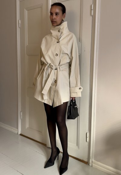 Mujer con gabardina beige con cinturón, medias negras transparentes, tacones altos negros puntiagudos, sosteniendo un pequeño bolso negro, de pie en el interior junto a la puerta.