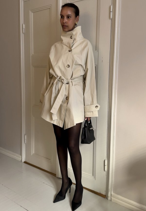 Kvinde iført beige trenchcoat med bælte, sorte gennemsigtige strømpebukser, sorte spidse høje hæle, holder en lille sort håndtaske, står indendørs ved døren.