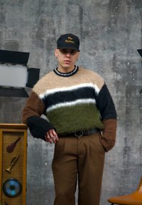 Pull en tricot à coupe oversize avec des rayures beige, vert, noir et blanc. Texture douce et duveteuse. Pantalon marron et casquette noire avec texte.