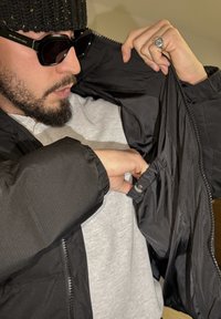 Veste noire légère avec une finition texturée, fermeture éclair et poignets élastiques. Comprend une poche intérieure accessible par l'avant.