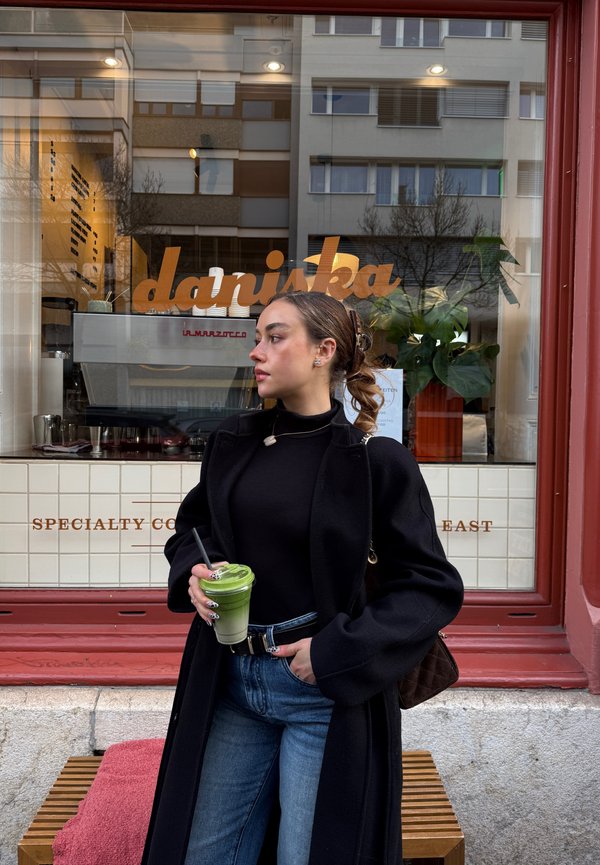 Jeune femme en manteau noir et jean tenant une boisson verte avec une paille, debout devant un café nommé "daniska" avec un banc à proximité.