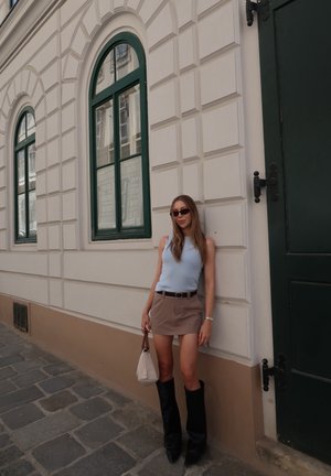 Joven mujer con gafas de sol, top sin mangas, minifalda y botas hasta la rodilla, se encuentra frente a una pared beige con ventanas arqueadas con marco verde.