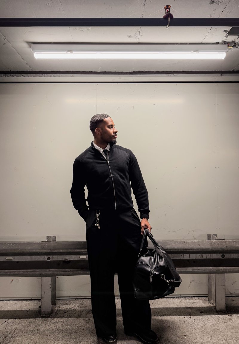 Homme en veste et pantalon noirs, debout, tenant un sac en cuir noir, regardant vers sa droite, devant un mur uni sous lumière fluorescente.