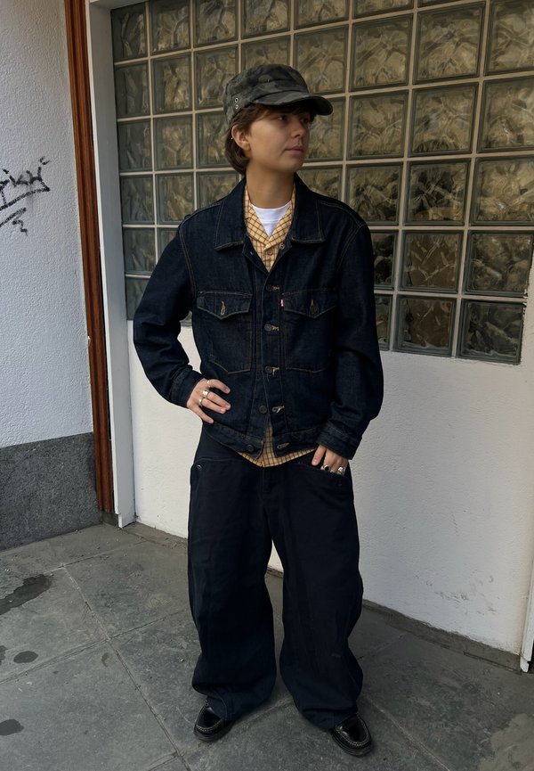 Person med mørk denimjakke, gul rutete skjorte, svarte vide bukser, svarte sko og en camo-caps som står mot en glassblokkvegg.