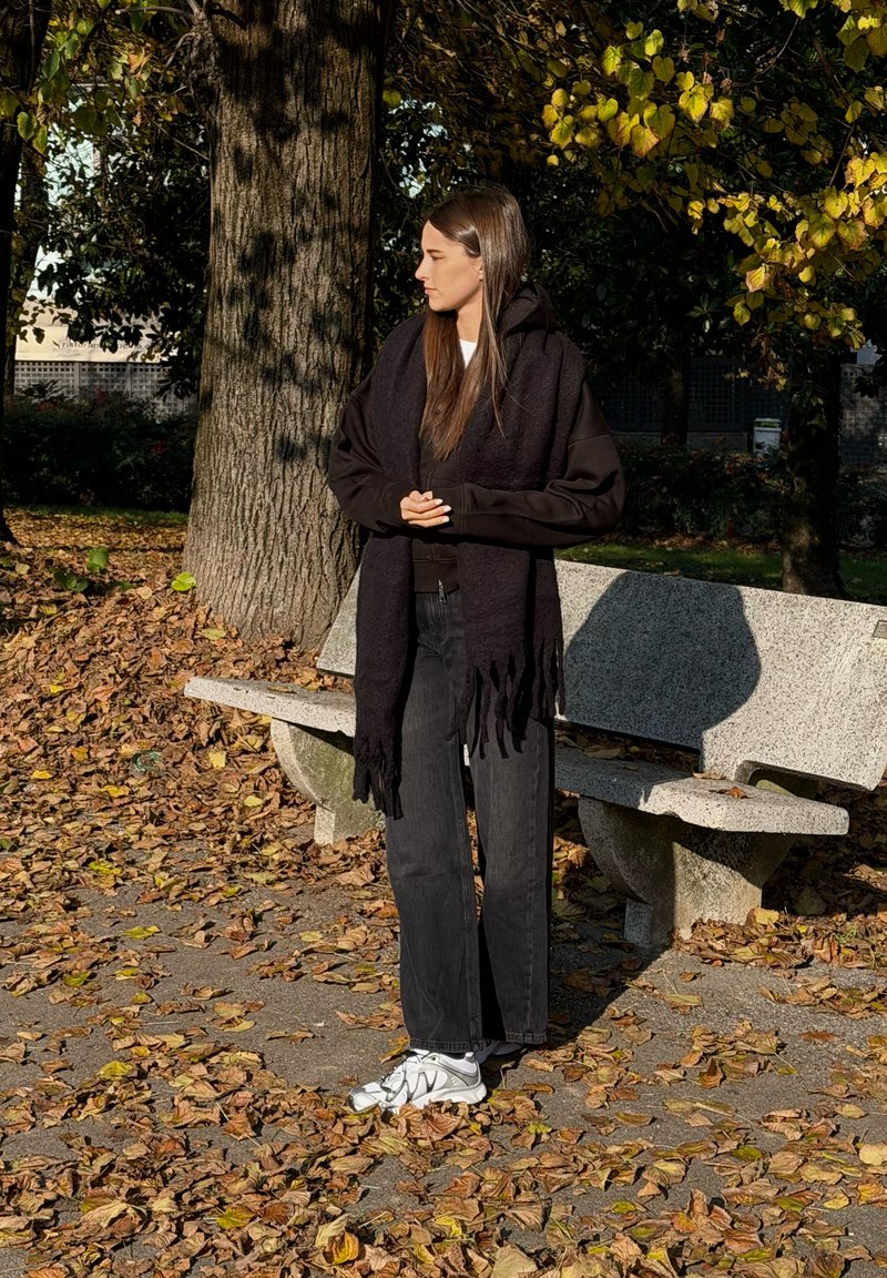 Xale preto usado sobre uma blusa preta, combinado com jeans soltos pretos e sapatilhas desportivas brancas. Em pé num caminho rodeado de folhas de outono.