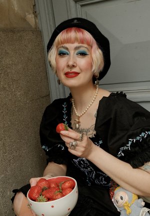 Un cuenco de fresas rojas frescas con verdes, sostenido por una persona que lleva un vestido negro bordado y accesorios, con un peluche cerca.