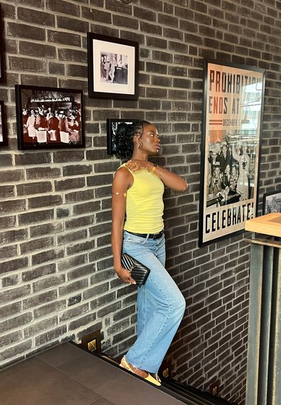 Top amarillo, jeans acampanados de un azul claro, bolso negro de mano. Fondo de pared de ladrillo con fotos en blanco y negro enmarcadas. Mostrador de madera al lado.