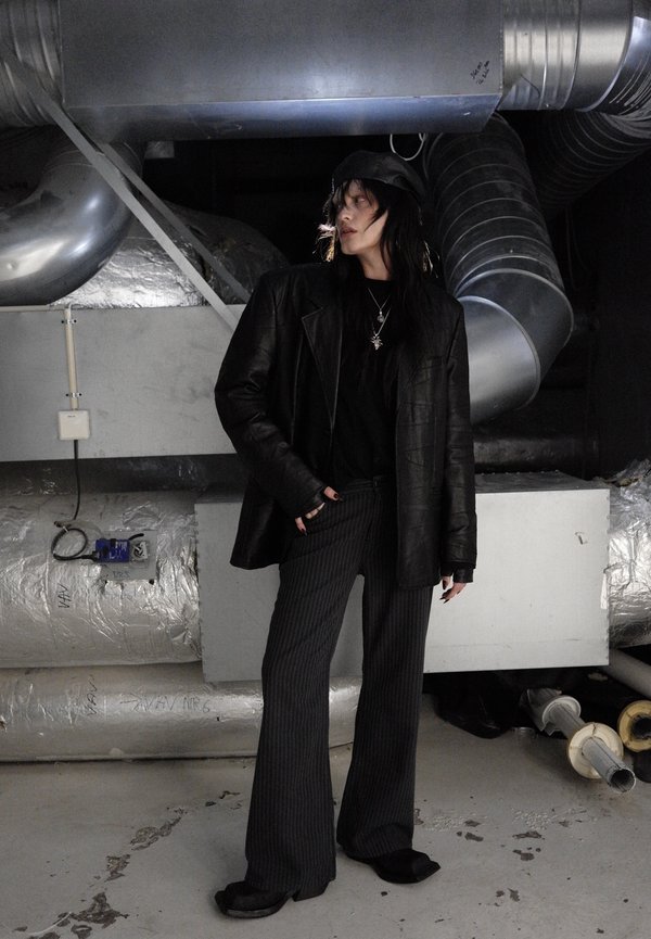 Persona con capelli lunghi e scuri che indossa una giacca di pelle nera, pantaloni a righe e un cappello, in piedi in un ambiente industriale con grandi tubi di metallo.
