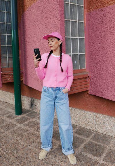 Cárdigan rosa suave, camisa blanca, vaqueros baggy azul claro y zapatos beige. Sosteniendo un teléfono inteligente negro, con pelo trenzado y una gorra rosa.