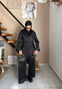 Μαύρο σατέν μακρύ παλτό με γιακά, σε συνδυασμό με φαρδιές μαύρες παντελόνες και χοντρές μαύρες πλατφόρμες. Αξεσουάρ: μικρή μαύρη τσάντα με αλυσίδα.