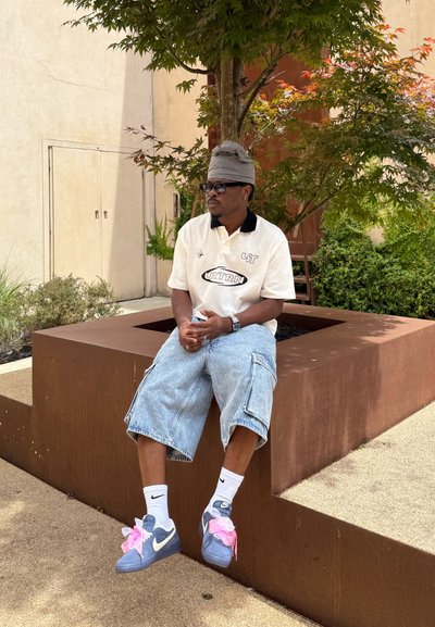 Persona vestida con una camisa polo blanca con collar negro, pantalones cortos de mezclilla tipo cargo y zapatillas Nike azules con acentos rosas, sentada en un macetero de concreto marrón.