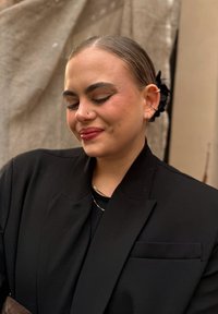 Black blazer with a pronounced shoulder line, styled with a black top and gold necklace. Complements natural makeup and a sleek updo.