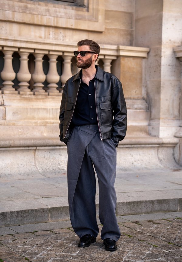 Homme portant une veste en cuir noire, une chemise foncée, un pantalon gris large et ample, des chaussures noires et des lunettes de soleil, debout à l'extérieur près d'une rambarde en pierre.