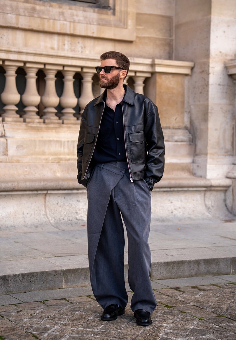Homme portant une veste en cuir noire, une chemise sombre, un pantalon gris ample à jambes larges, des chaussures noires et des lunettes de soleil, debout à l'extérieur près d'une rambarde en pierre.