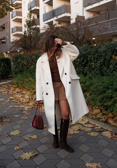 Abrigo de lana blanco de gran tamaño con botones negros, suéter marrón de canalé, falda color crema, botas marrones hasta la rodilla y un bolso rojo. Hojas de otoño en el fondo.