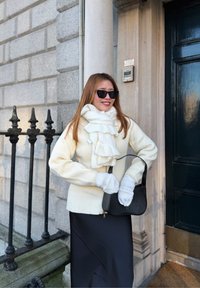 Femme portant une écharpe blanche, des gants et un pull avec une jupe noire et des lunettes de soleil, tenant un sac à main noir devant l'entrée d'un bâtiment en pierre.
