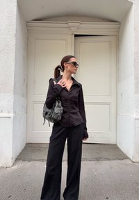 Camisa preta de botão com detalhe franzido, mangas longas; combinada com calças largas pretas; segurando uma bolsa preta texturizada com tachas.