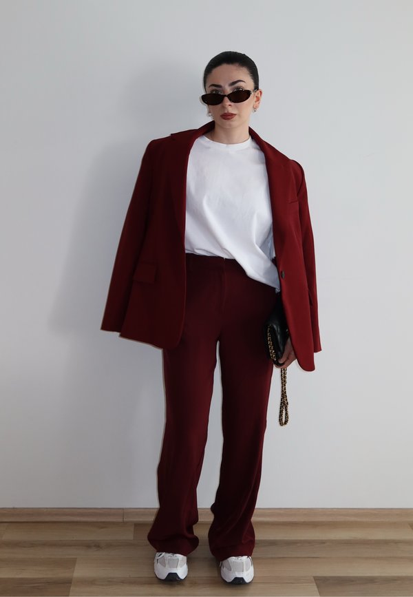 Femme portant un costume rouge foncé avec blazer posé sur les épaules, t-shirt blanc, baskets blanches, lunettes de soleil noires, et tenant un sac noir avec chaîne dorée.