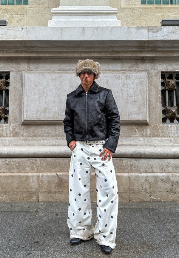 Persona con giacca nera, pantaloni bianchi a gamba larga con pois neri, cappello di pelliccia, occhiali da sole, numerosi anelli, in piedi contro un muro di pietra.