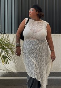 Kleid aus Spitze in Weiß mit einem geschichteten Design und Rüschensäumen, kombiniert mit dunklen Hosen. Die Accessoires umfassen eine schwarze Kappe und silbernen Schmuck.