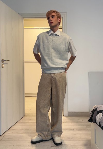 Suéter sin mangas gris claro sobre una camisa blanca con cuello, combinado con jeans beige de pierna ancha y zapatos blancos. Diseño minimalista y tonos neutros.