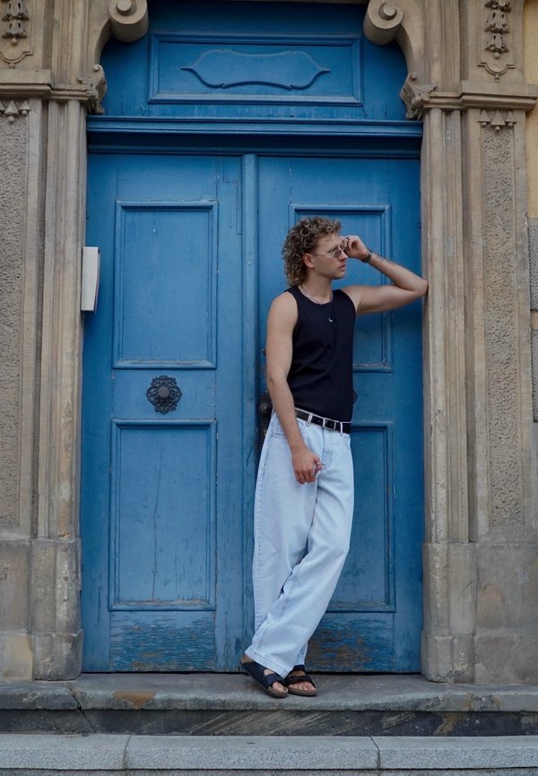 Top preto sem mangas, jeans soltos de cor azul clara e sandálias pretas. A pessoa apoia-se contra uma porta de madeira azul com detalhes ornamentais.