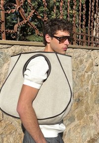 Siva torbica za nošenje z rjavimi poudarki, s krožnimi izrezi za položaj rok. Nošena ob kamniti steni kot ozadju.