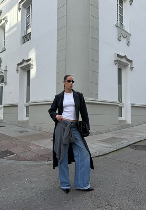 Mujer de pie en la esquina de la calle, vistiendo un abrigo largo negro, blusa blanca, vaqueros anchos, gafas de sol y sosteniendo una bolsa negra.