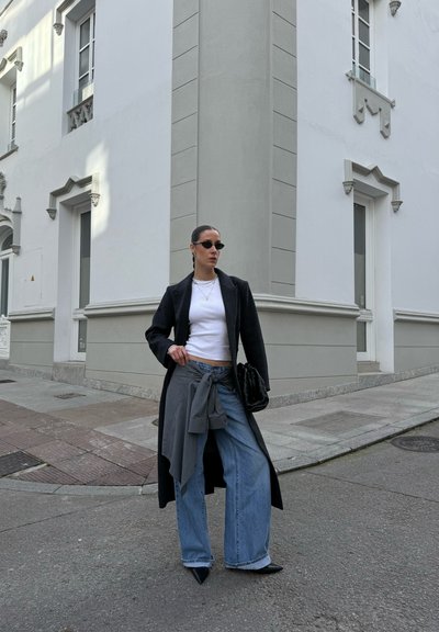 Mujer de pie en la esquina de la calle, vistiendo un abrigo largo negro, blusa blanca, vaqueros anchos, gafas de sol y sosteniendo una bolsa negra.