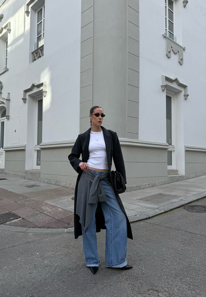 Femme debout à un coin de rue, portant un long manteau noir, un haut blanc, un jean large, des lunettes de soleil, et tenant un sac noir.
